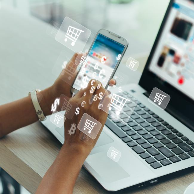 A person online shopping on their phone and laptop. Photo is overlaid with dollar signs and shopping cart icons.